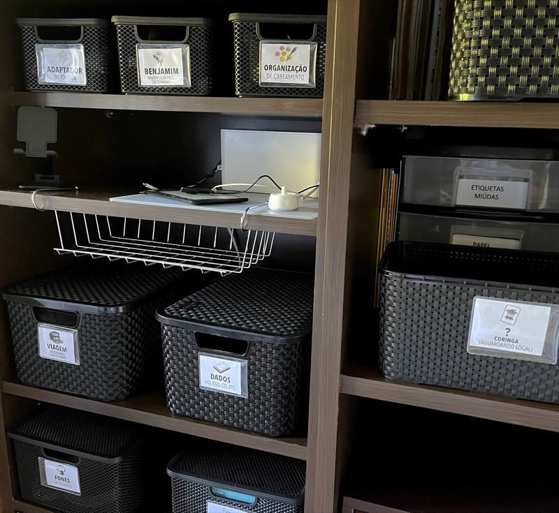 Foto de algumas prateleiras cheias de caixas pretas, cada uma com etiqueta descrevendo seu conteúdo: organização de cabeamento, adaptadores de tomadas, fones de ouvido, etc.