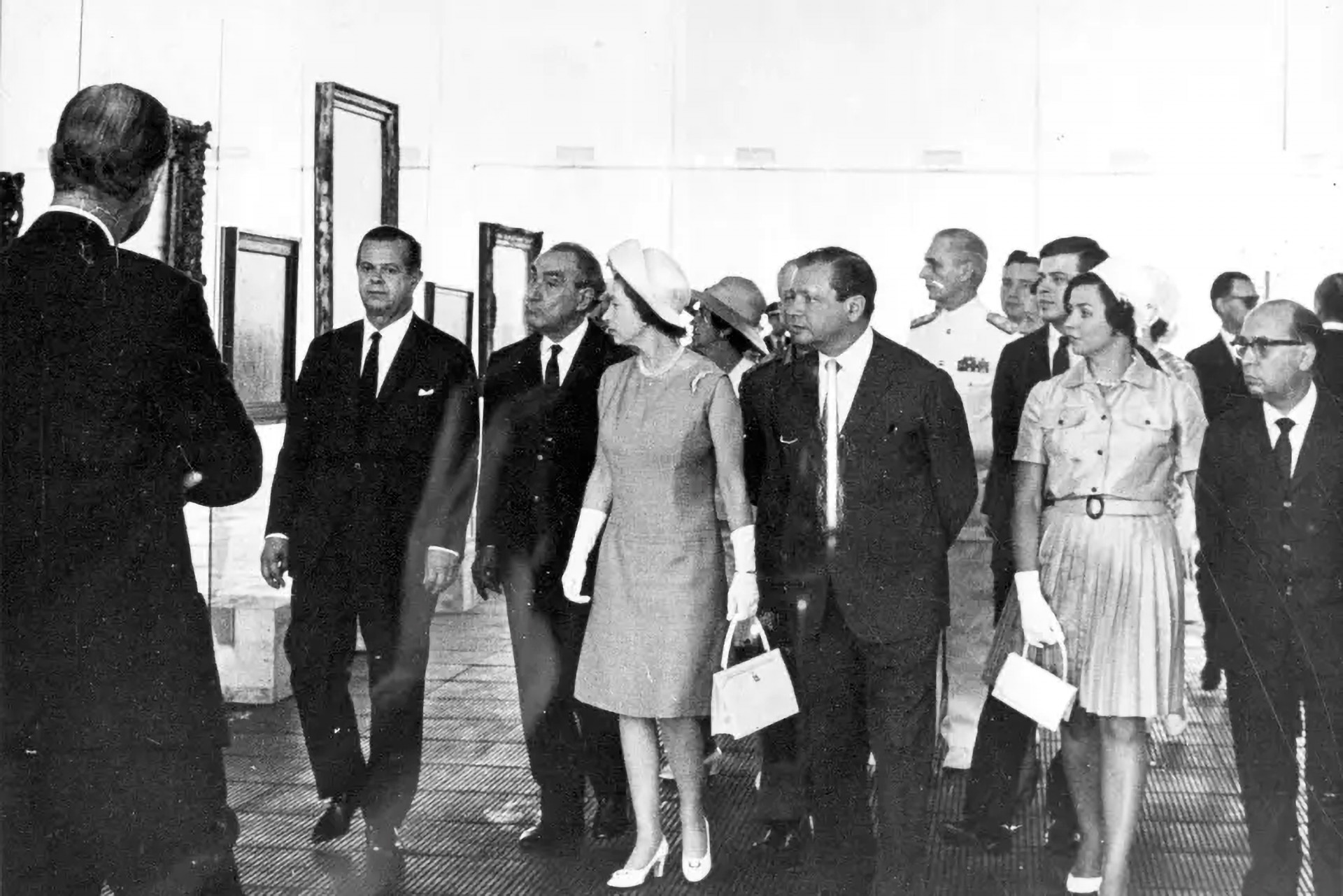 A rainha Elizabeth II observa as pinturas em exposição, cercada por uma entourage de autoridades.