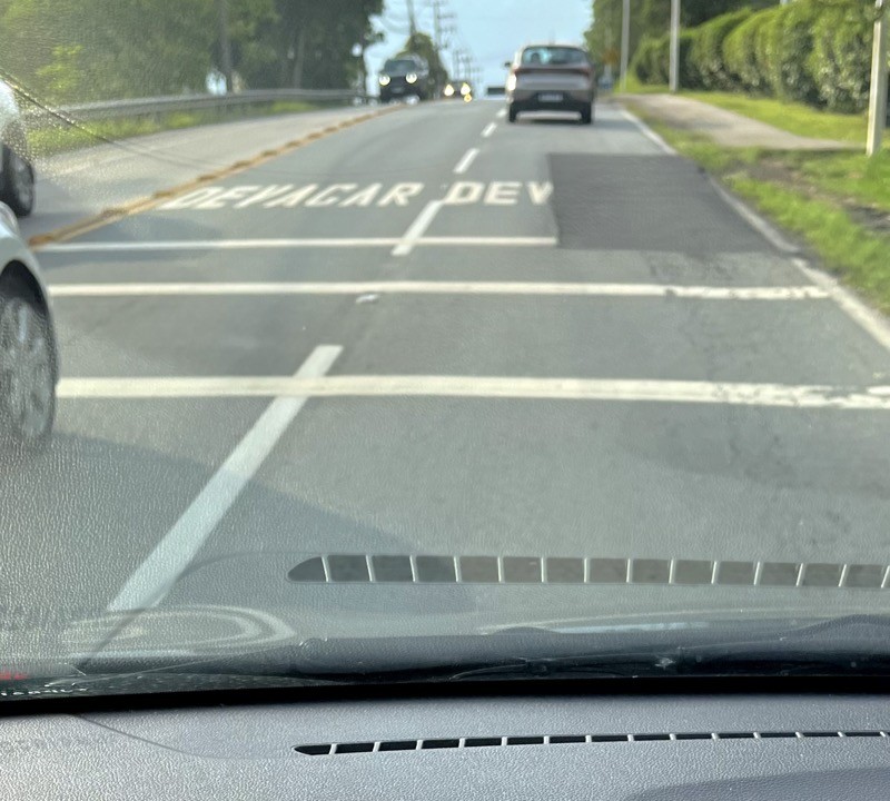Fotografia do asfalto da rodovia SC-404, em trecho onde havia o alerta “DEVAGAR“ escrito em cada uma das duas pistas do sentido bairro-centro, mas uma obra com recapeamento asfáltico removeu as 4 últimas letras do alerta em uma das pistas.