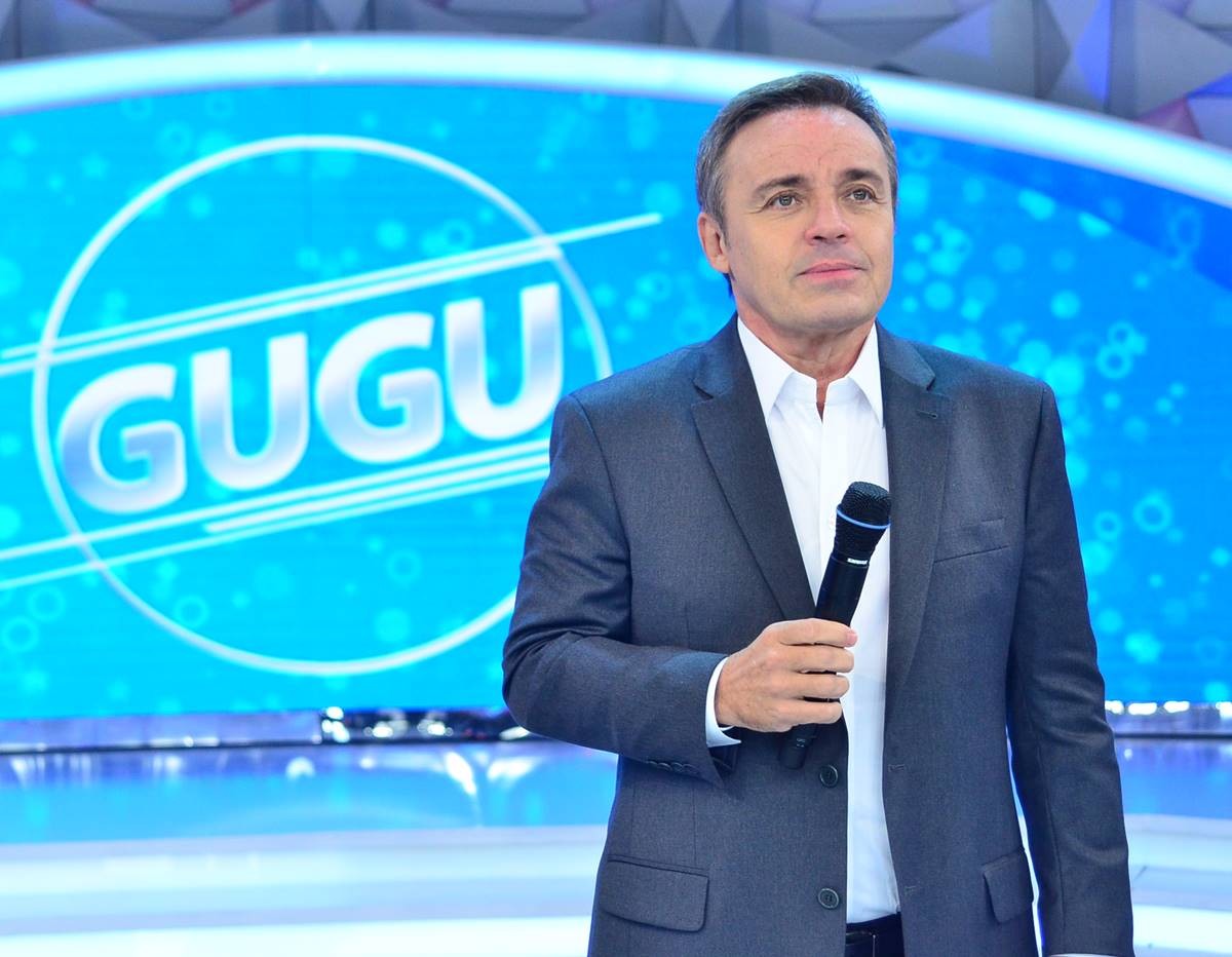 Gugu Liberato olha para o alto, com microfone em punho, no cenário de seu programa