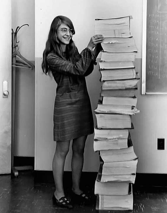 Margaret Hamilton sorri junto à pilha de 17 volumes de código-fonte impresso, da programação do computador de bordo da Apollo 11.<br>