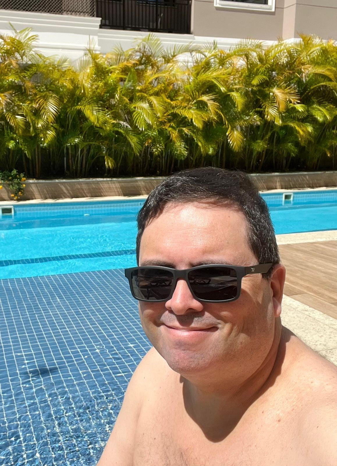 Foto minha, de óculos escuros, olhando para a câmera e sorrindo, em ambiente de piscina cercado por vegetação ornamental