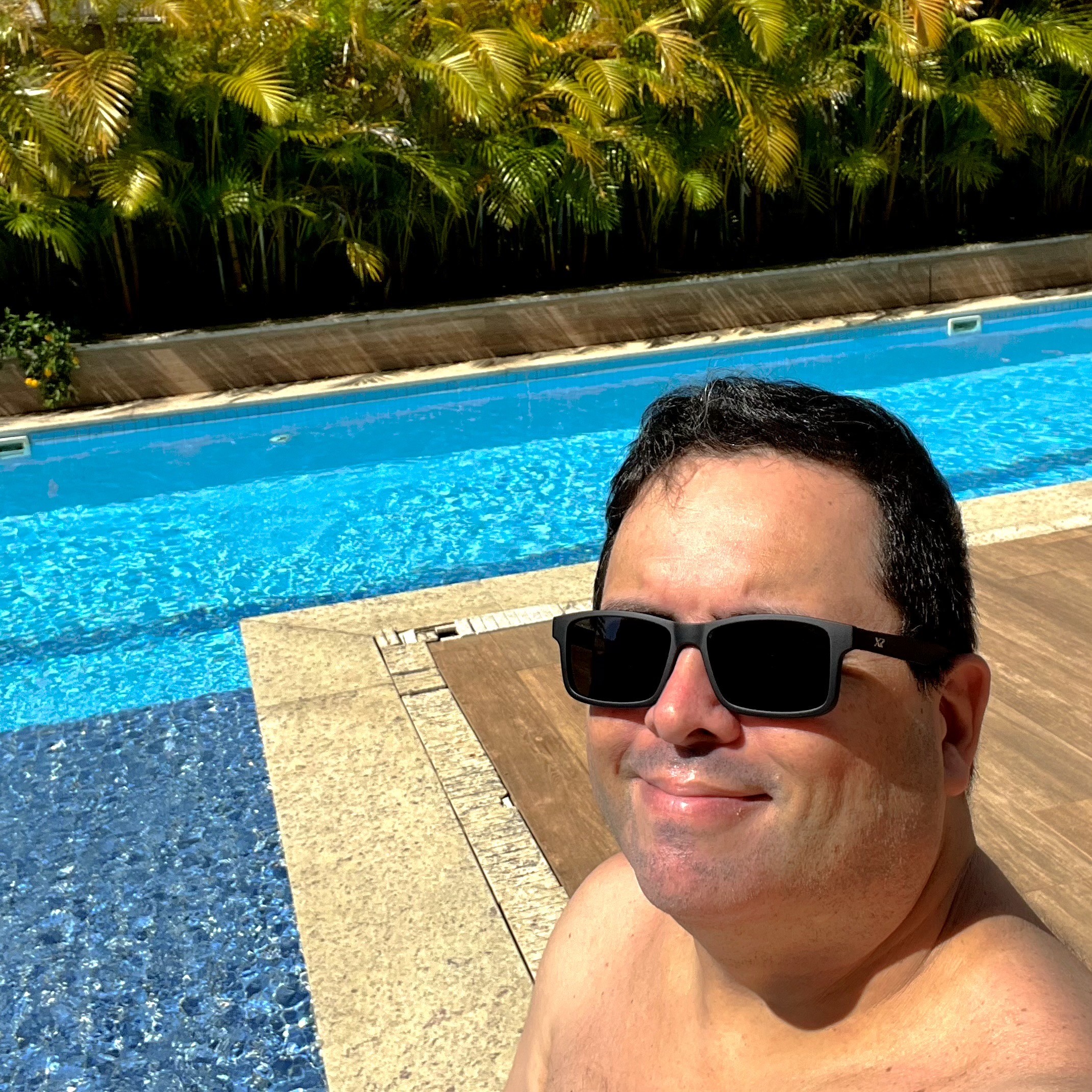 Foto minha, sem camisa e de oculos escuros, sorrindo para a câmera em ambiente ensolarado da piscina