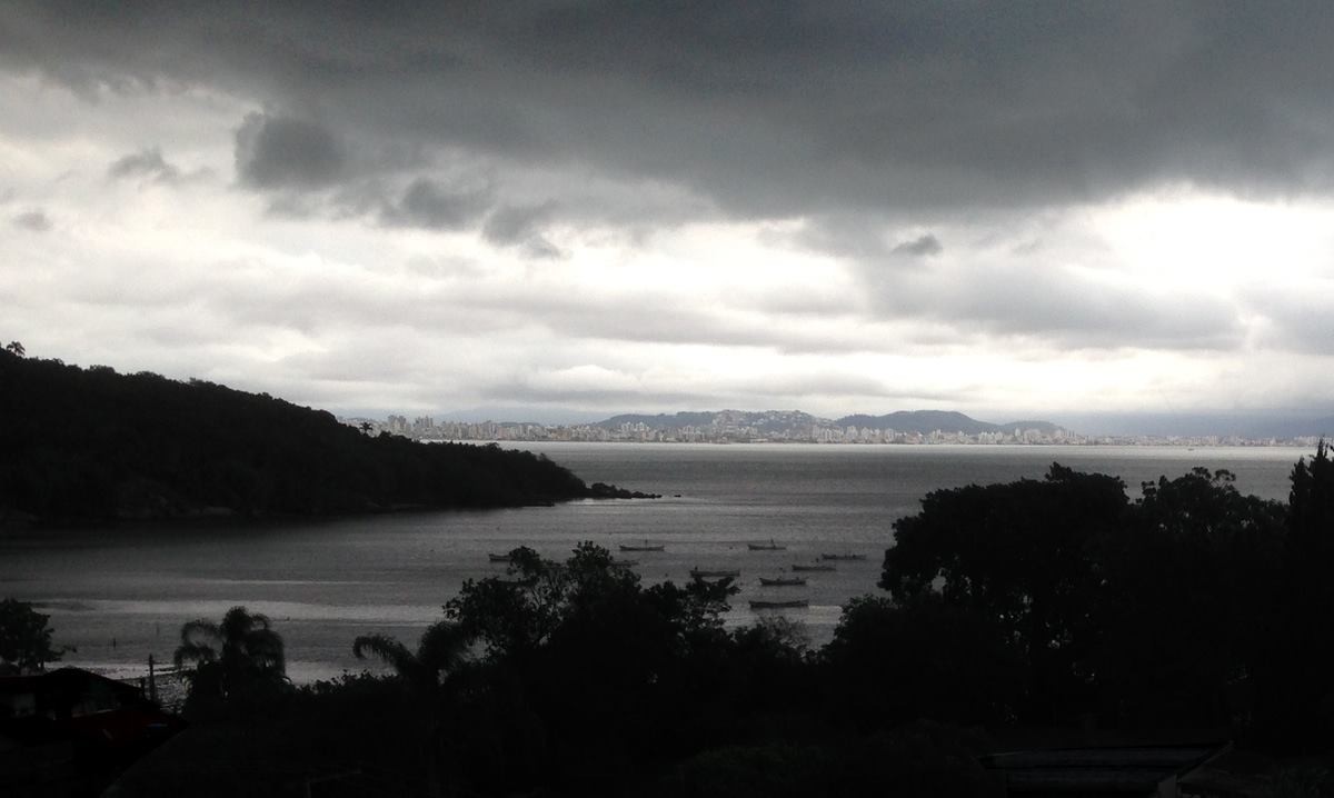 Vista do porto de pescadores do bairro João Paulo, com várias embarcações de pequeno porte abrigadas, sob um céu bem escuro. À distância, vê-se o centro de Biguaçu, um município vizinho.