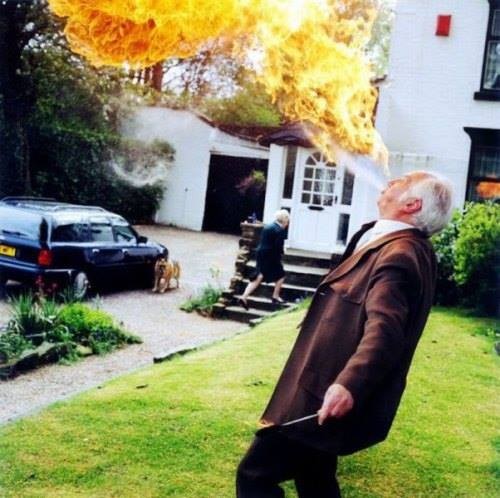 Um homem grisalho, na frente de casa, de paletó e gravata, cospe uma grande bola de fogo, enquanto o cachorro observa e a uma senhora caminha na direção oposta