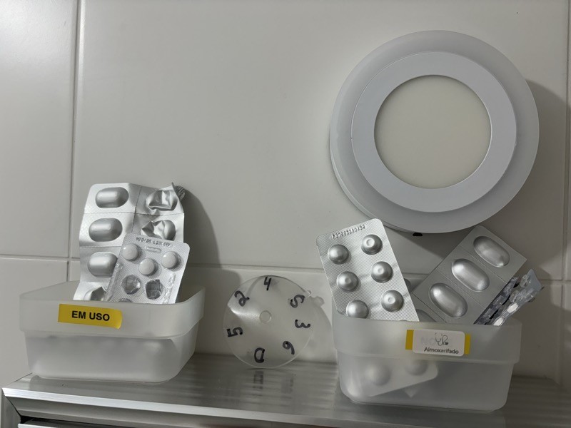 Um disco de plástico com indicativos dos 7 dias da semana, escritos com marcador permanente, posicionado entre 2 caixas com embalagens de comprimidos, com etiquetas “em uso“ e “almoxarifado“