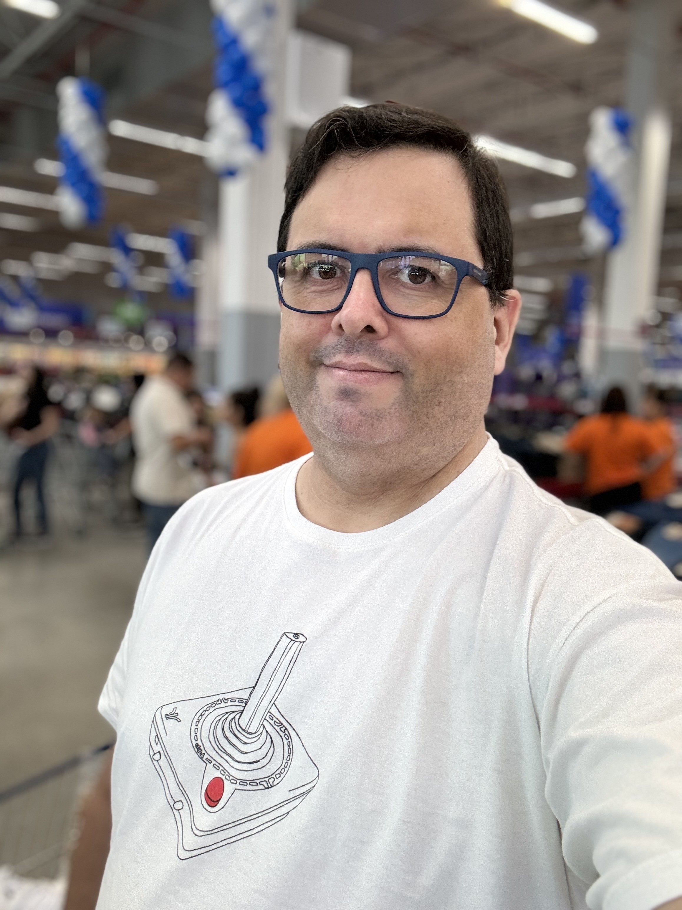 Foto minha durante as compras de supermercado, olhando para a câmera, com camiseta mostrando um desenho de joystick de Atari 2600