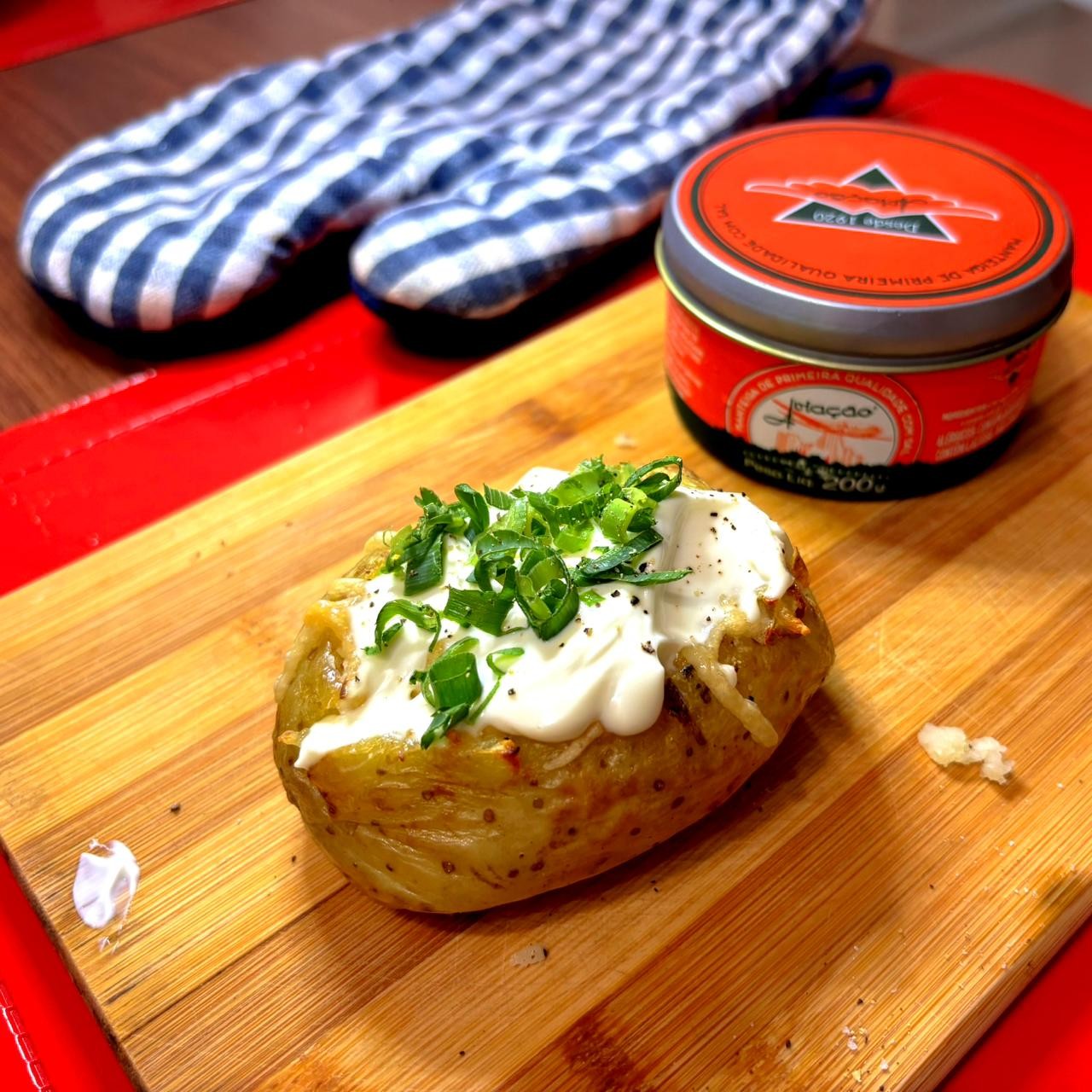 Uma batata assada recheada, coberta com requeijão e com cebolinha picada, colocada sobre uma tábua de corte, ao lado de uma lata de manteiga e de uma luva de manipular panelas
