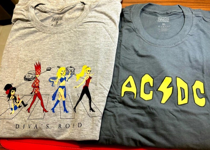 Uma camiseta cinza com estampa similar à capa do disco Abbey Road, só que ao invés dos Beatles, quem atravessa na faixa são: Madonna, Britney, Lady Gaga e Amy Winehouse. Ao lado, uma camiseta cinza estampada com a sigla AC/DC em amarelo.
