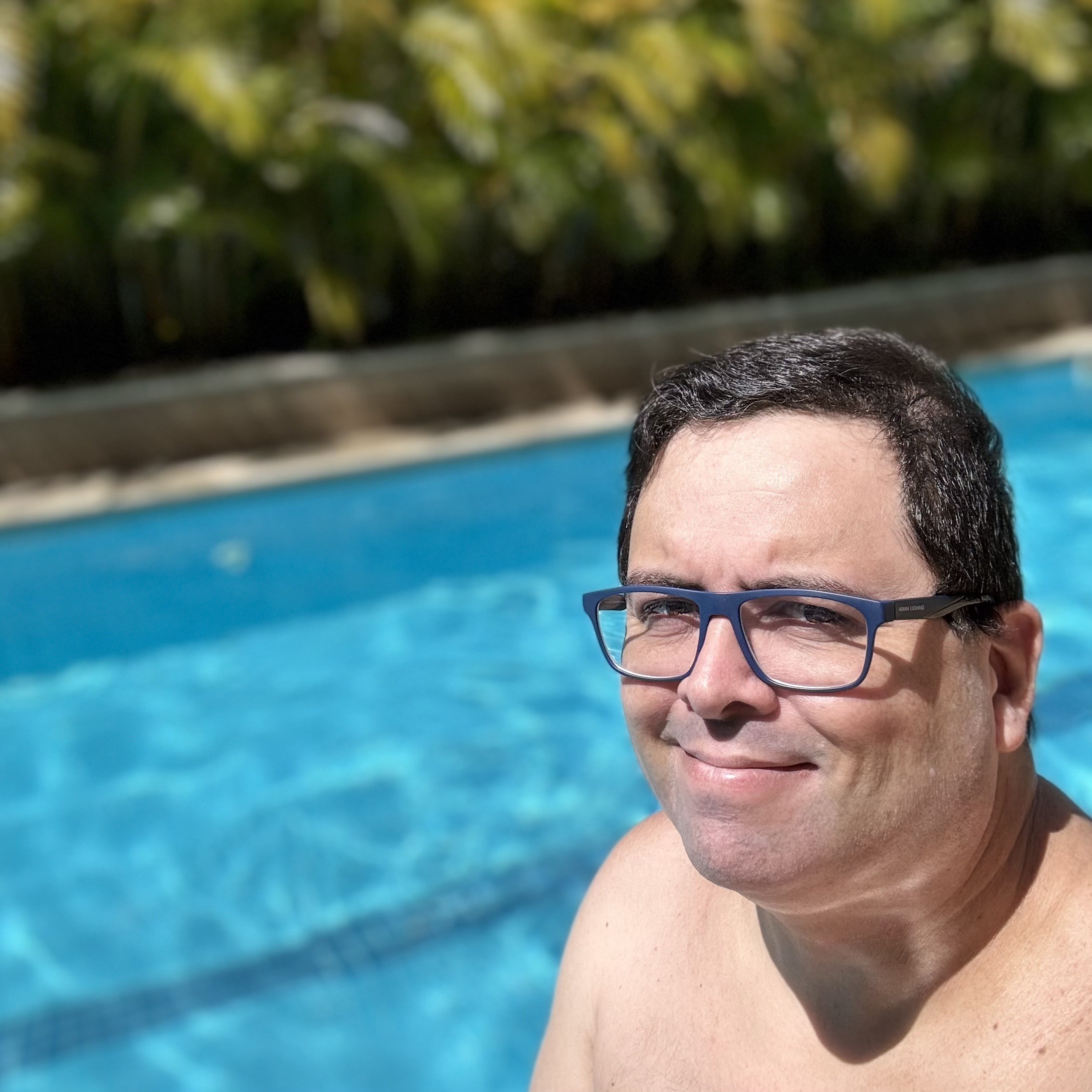 Foto minha olhando para a câmera e sorrindo, de oculos e sem camisa, em ambiente de piscina, ensolarado 