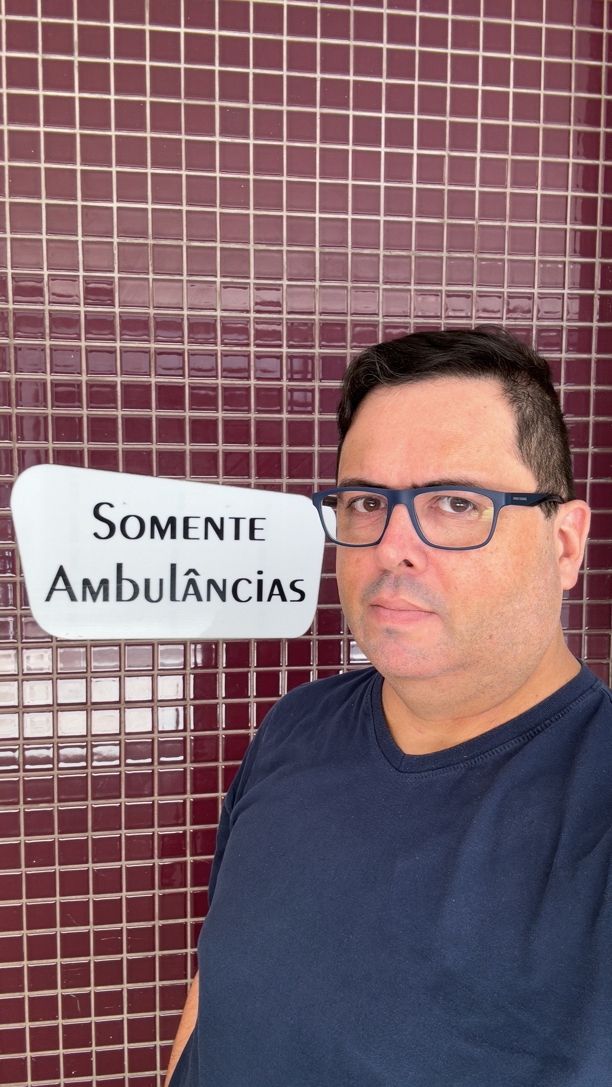 Eu, de camisa azul e oculos de armação azul, olho para a câmera, ao lado de um cartaz onde se lê: SOMENTE AMBULÂNCIAS