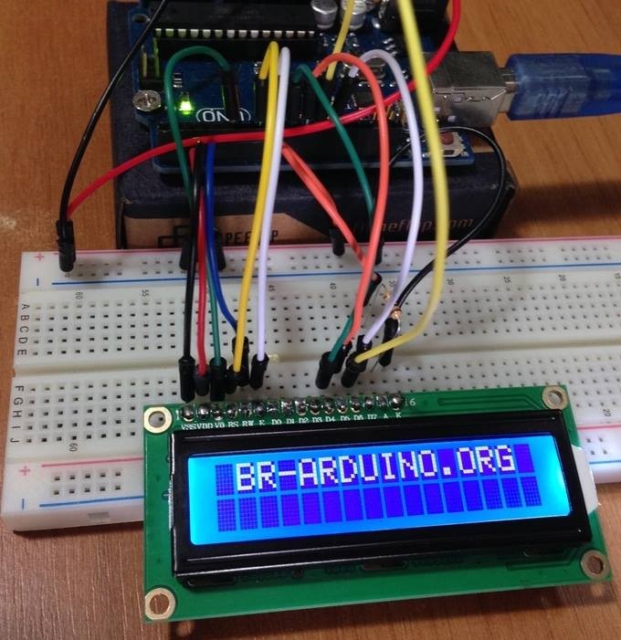 Um display LCD de duas linhas e 16 caracteres em cada linha, exibindo o nome do meu site br-arduino.org, conectado a uma breadboard com vários jumpers coloridos, conectados a uma placa Arduino Uno.