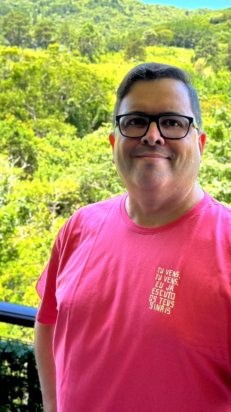 Foto minha olhando para a câmera, com a mata ensolarada ao fundo, usando uma camiseta em tom de rosa com um trecho de música escrito na altura do peito: tu vens, tu vens, eu já escuto teus sinais