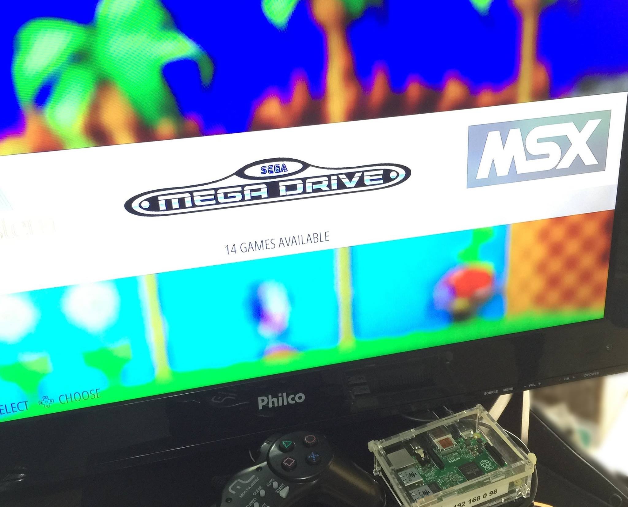 Tela de uma TV mostrando um menu para escolher entre 14 jogos do Mega Drive e do MSX. Abaixo dela podem ser vistos um controle Multilaser e um raspberry pi com uma etiqueta indicando o seu endereço IP, colada em um case transparente
