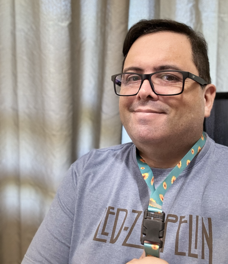 Eu, com camiseta do Led Zeppelin e óculos de armação azul, sorrio para a câmera, segurando um cordão para crachá em cor verde e com desenho de girassóis, da Lei 14.624, que formaliza seu uso para identificar pessoas com deficiências ou transtornos ocultos.