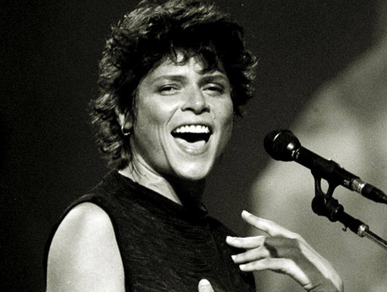 Em foto em preto e branco, Cássia ELler sorri para a câmera, em frente ao microfone de pedestal.