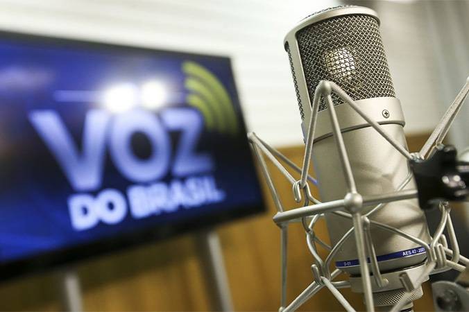 Microfone de estúdio de rádio, com um banner da Voz do Brasil ao fundo.