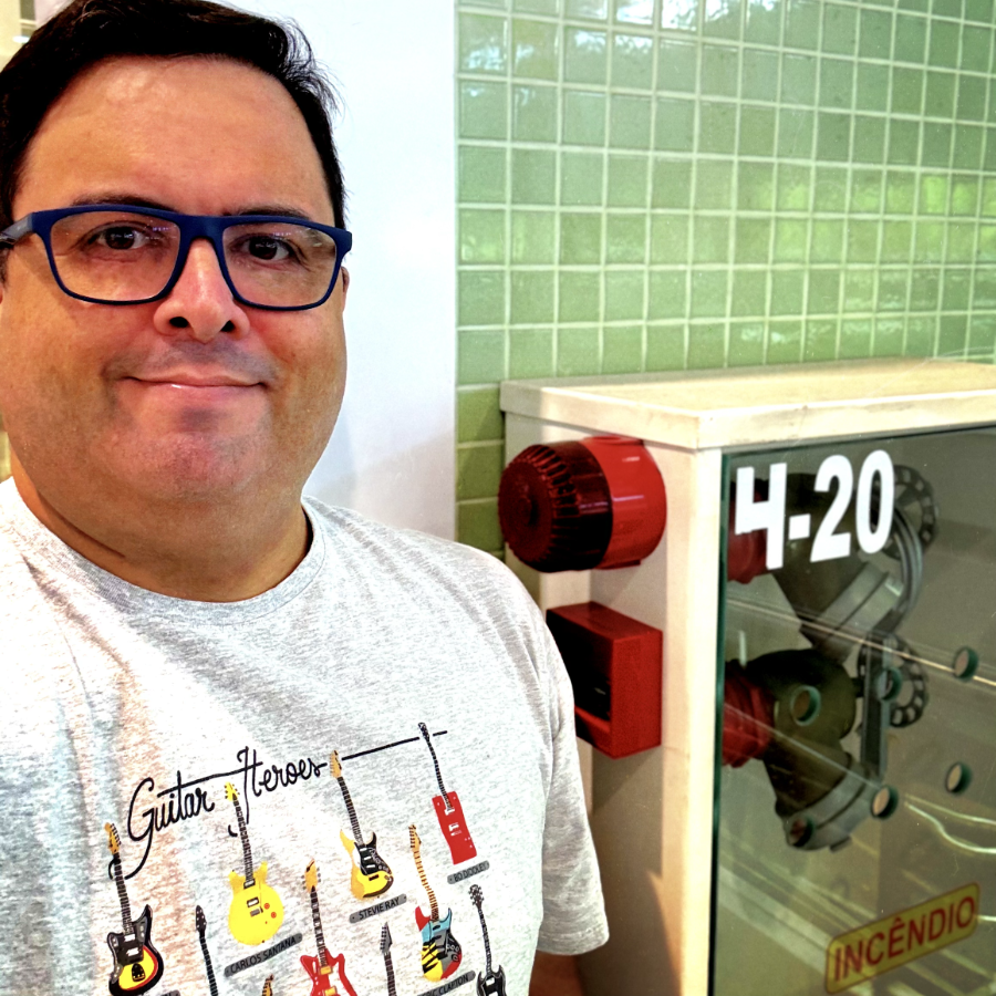 Foto minha olhando para a câmera, de óculos de armação azul e camiseta cinza com o título ‘Guitar Heroes’, mostrando várias guitarras célebres, incluindo as de Santana, Stevie Rai Vaughan e Bo Diddley. Ao meu lado está um armário de incêndio, com sirene, e a identificação 4-20.