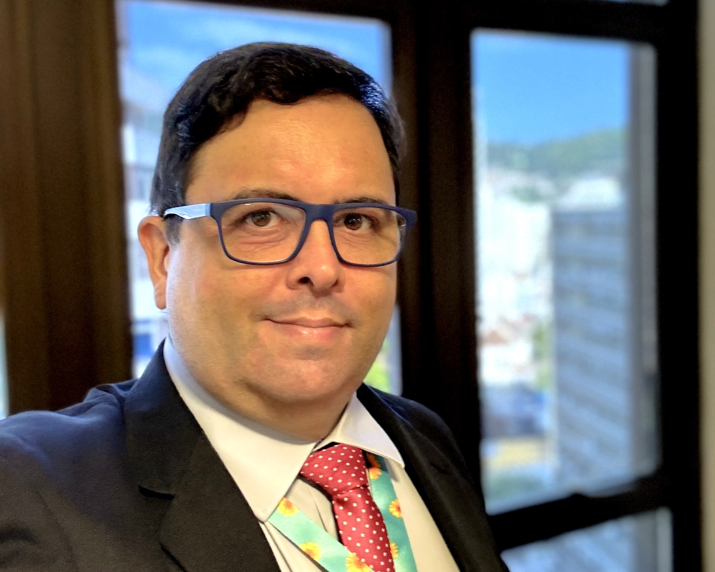 Eu olhando para a câmera, de óculos azul, paletó escuro, camisa clara, gravata vermelha e cordão de girassol. Ao fundo, paisagem urbana desfocada. 