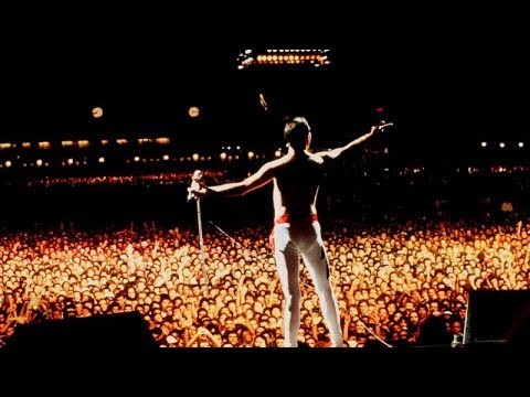 De costas, visto do palco, Freddie Mercury atua como regente da plateia