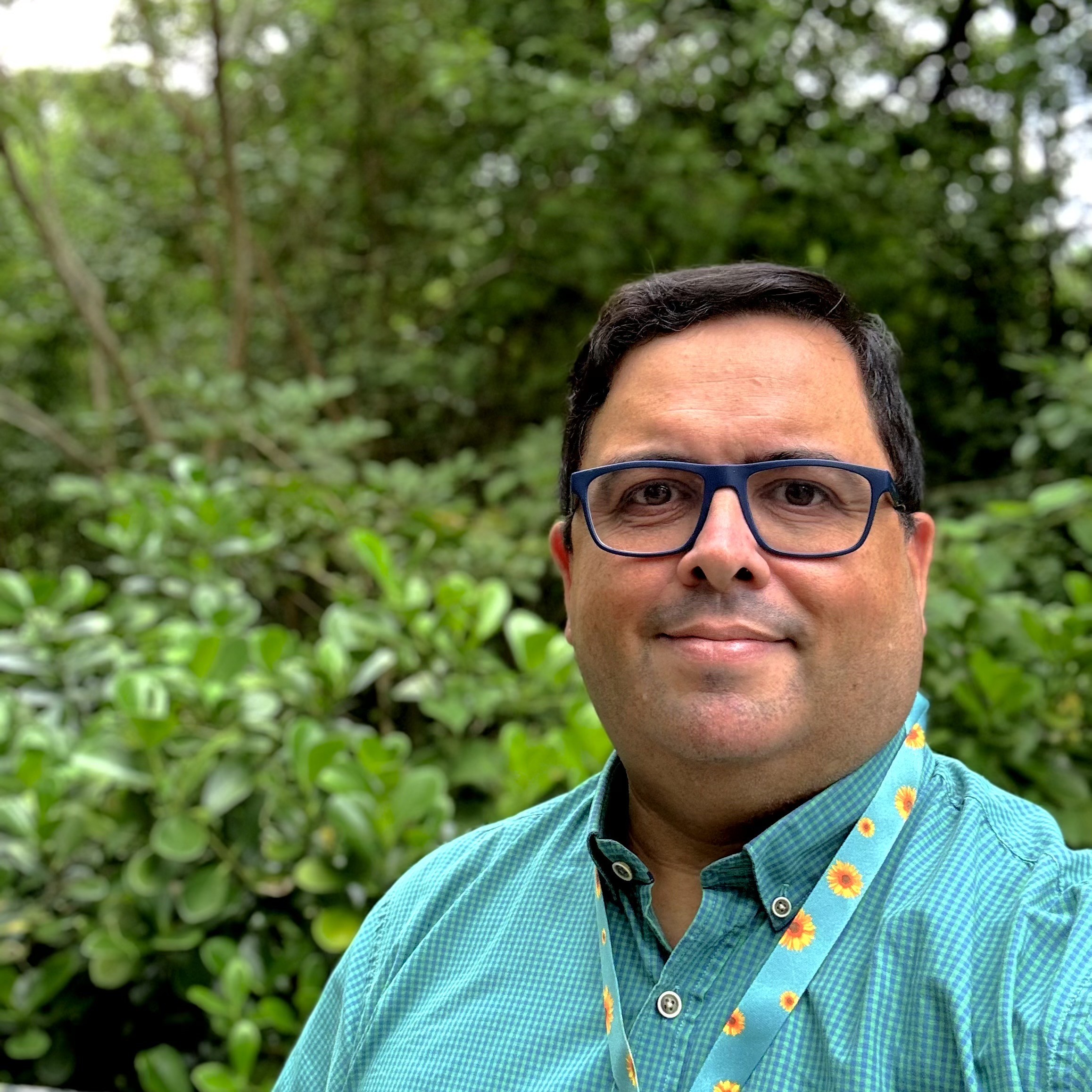 Foto minha olhando para a câmera, de camisa verde e usando um cordão ao redor do pescoço, com estampa de girassol. Ao fundo a vegetação. 