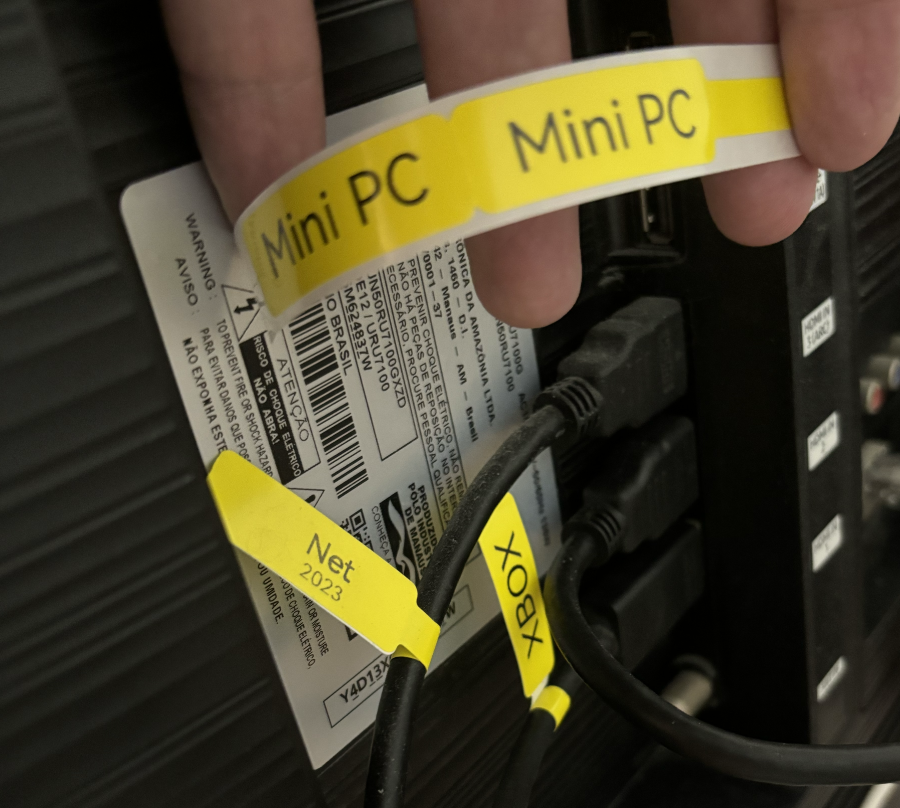 Dois cabos HDMI identificados com etiquetas amarelas que dizem “Net“ e Xbox“, e uma mão segurando uma etiqueta pronta para ser aplicada, onde se lê “Mini PC“