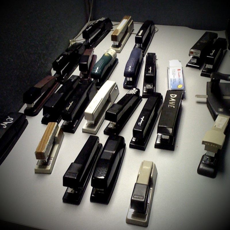 Foto de 23 grampeadores, de variados modelos, sobre o tampo de uma mesa de escritório