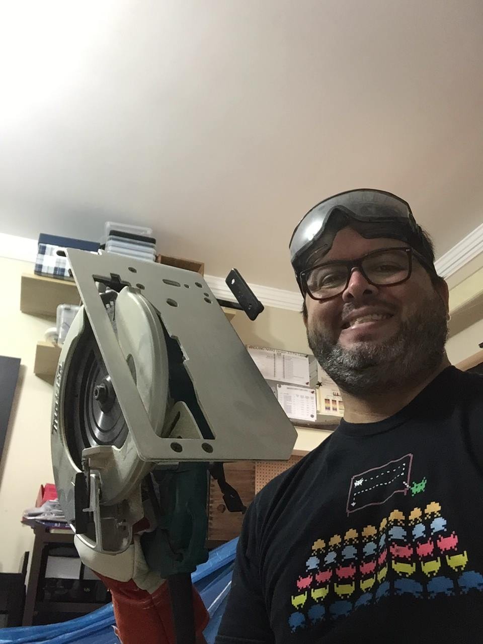 Foto minha, de barba e óculos (e com um óculos de proteção temporariamente apoiado na testa), segurando uma serra circular e com um largo sorriso no rosto, na oficina, vestindo uma camiseta mostrando a academia de combate dos Space Invaders do jogo de mesmo nome.