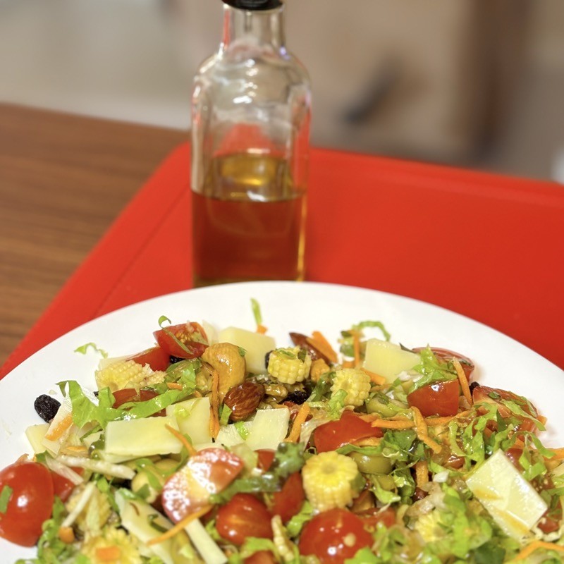Meu prato de salada contendo folhas verdes picadas, tomate picado, queijo picado, minimilho picado, mix de castanhas, azeitonas picadas. Ao fundo, a garrafa de azeite de oliva.