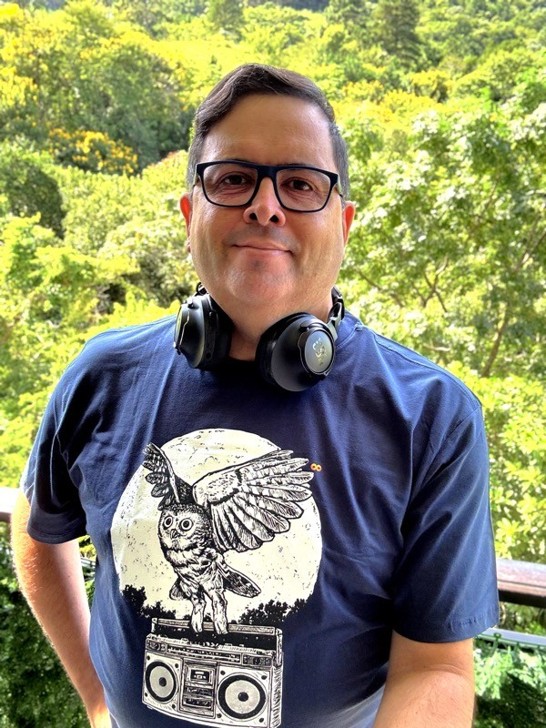 Foto minha olhando para a câmera, com a mata ensolarada ao fundo, com camiseta em que uma coruja voa levando consigo um aparelho de som com 2 falantes e tape-deck.