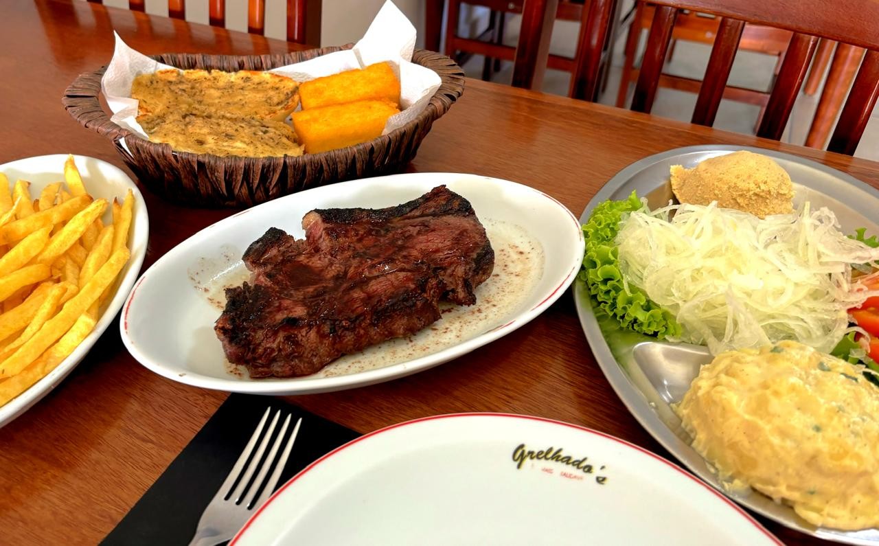 mesa com travessas contendo um assado de carne, polenta, batata frita, maionese, farofa e salada