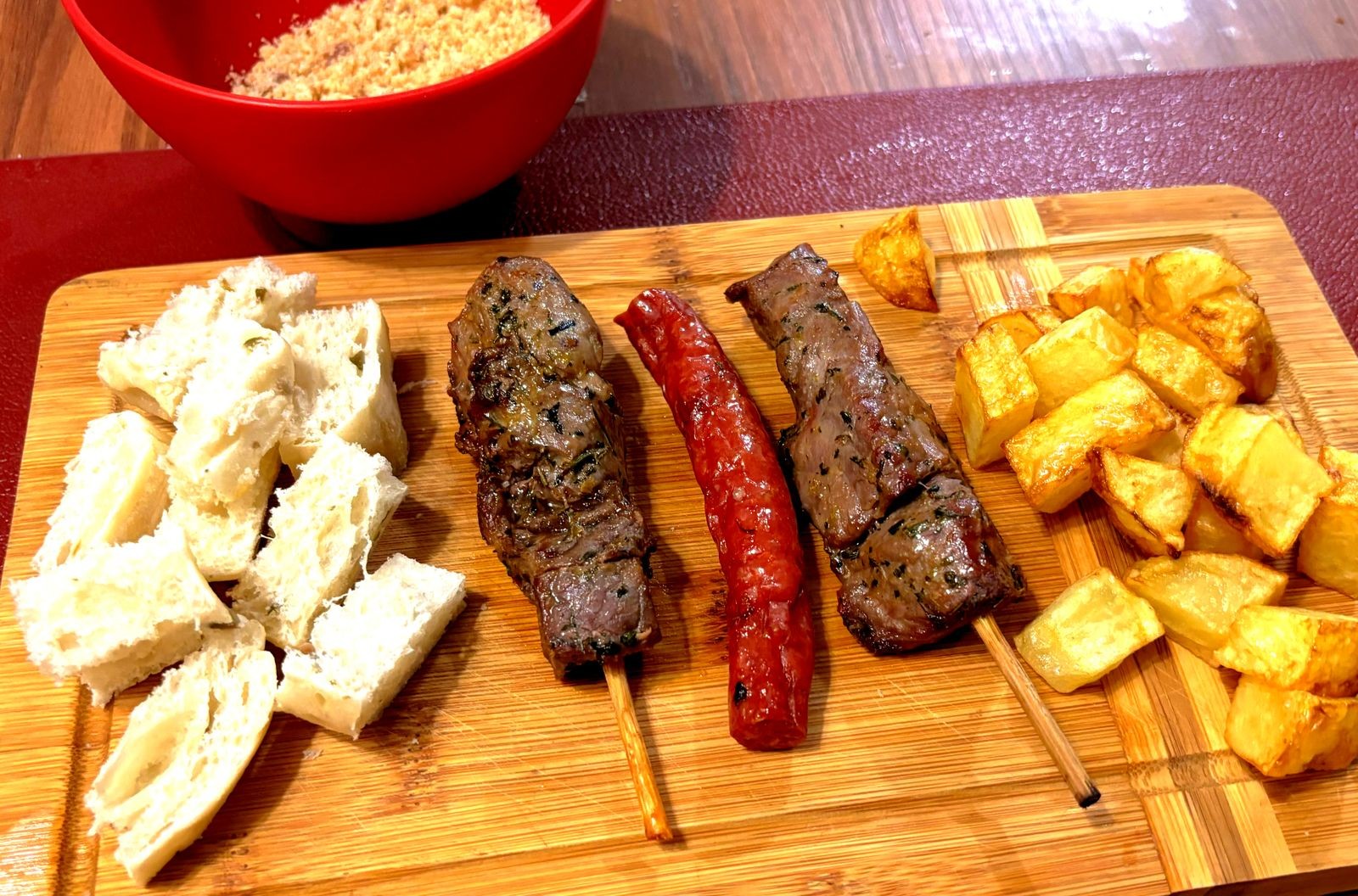 uma tábua contendo cubinhos de pão, 2 espetinhos de contrafilé, uma linguicinha e algumas batatas fritas cortadas em forma aproximada de cubos rombóides, ao lado de uma travessa de farofa