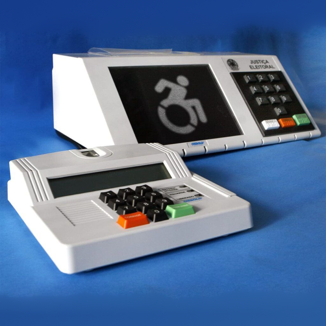 Foto de um terminal do mesário e de uma urna eletrônica, com um stencil do símbolo internacional da acessibilidade aplicado à tela da urna.