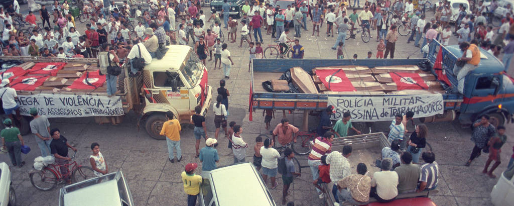 Dois caminhões carregados de caixões chegam à cidade em frente à população reunida.