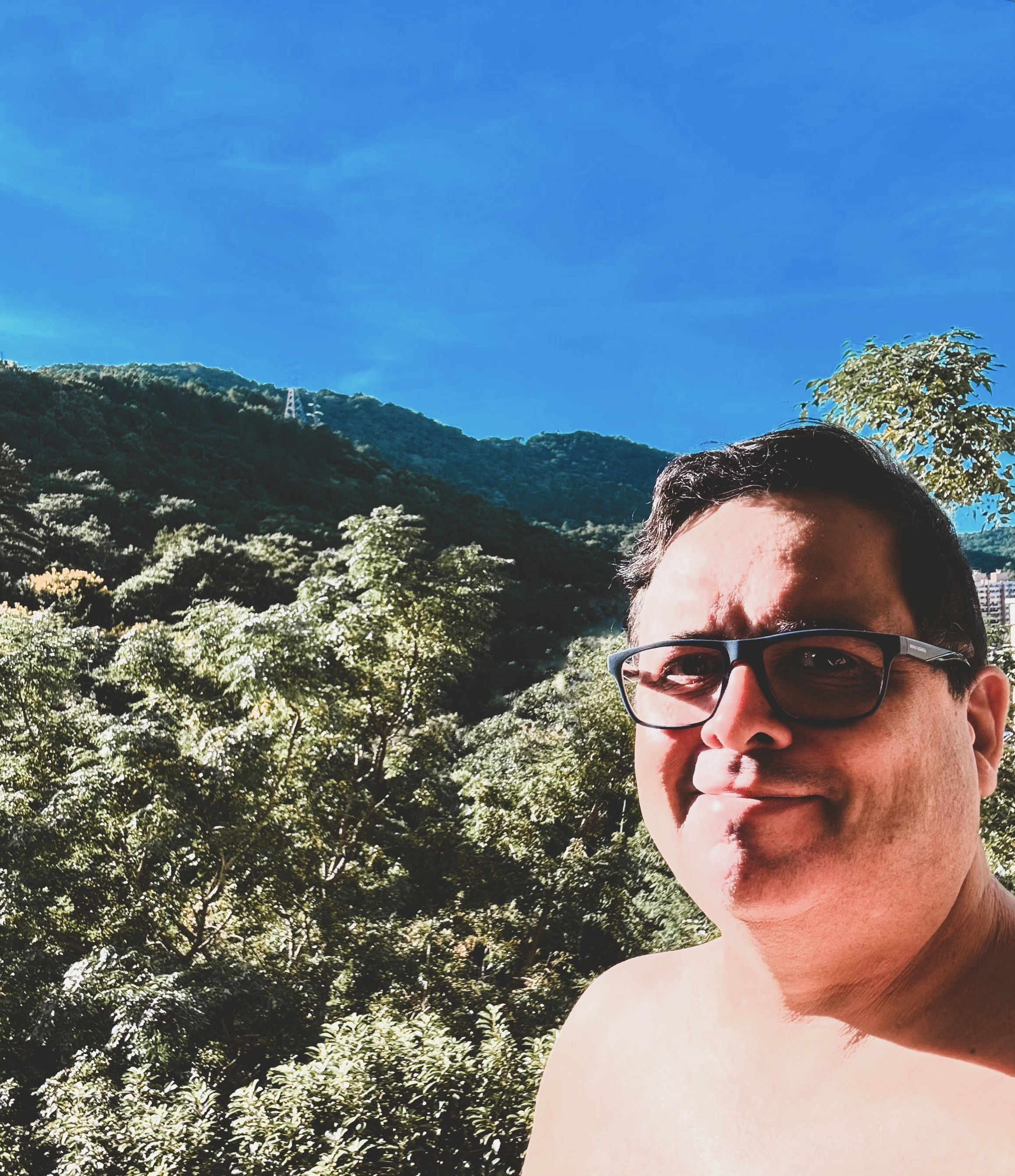 Foto minha sem camisa, olhando para a câmera,  com a mata e o céu azul ao fundo. 