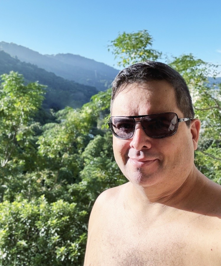 Foto minha, de óculos escuros e sem camisa, olhando para a câmera, com a mata, os morros e o céu azul ao fundo