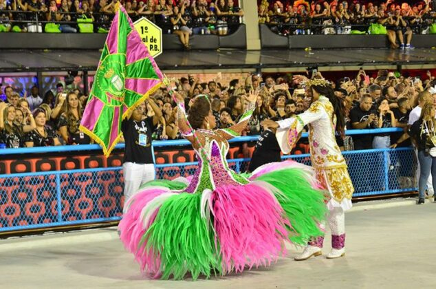 Porta bandeira com fantasia nas cores verde e rosa da escola, e seu mestre-sala.