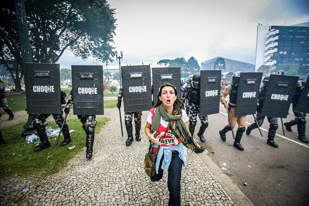 Foto de mulher grevista correndo e, atrás dela, a linha da PM de Choque avança, com escudos e cassetetes.