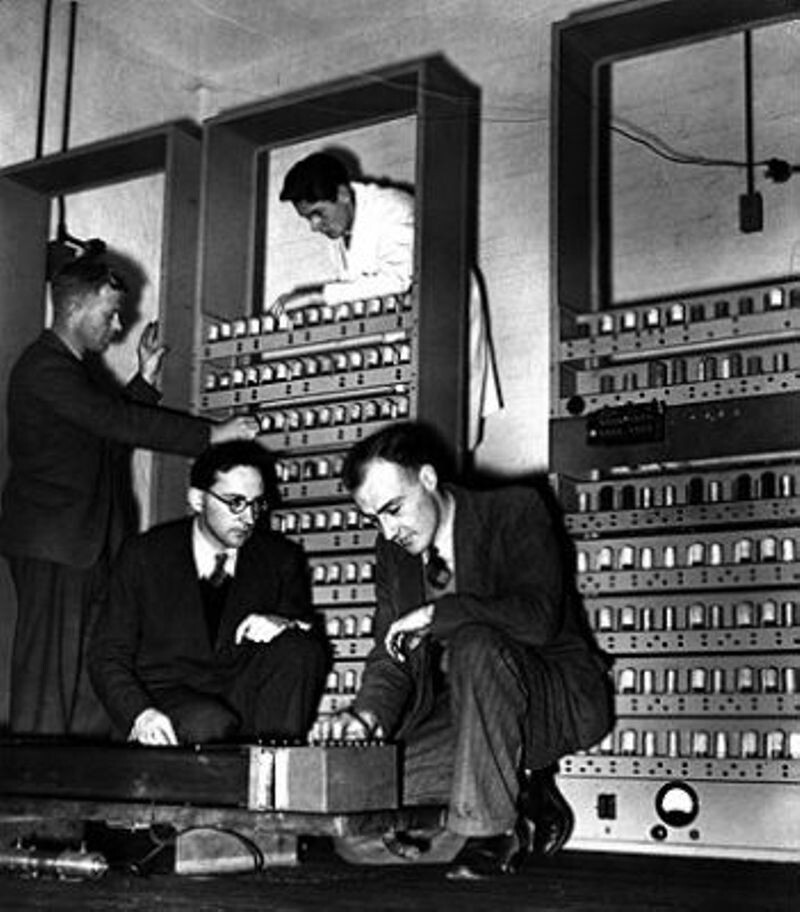 Foto em preto e branco de 3 engravatados e um portador de jaleco montando o EDSAC