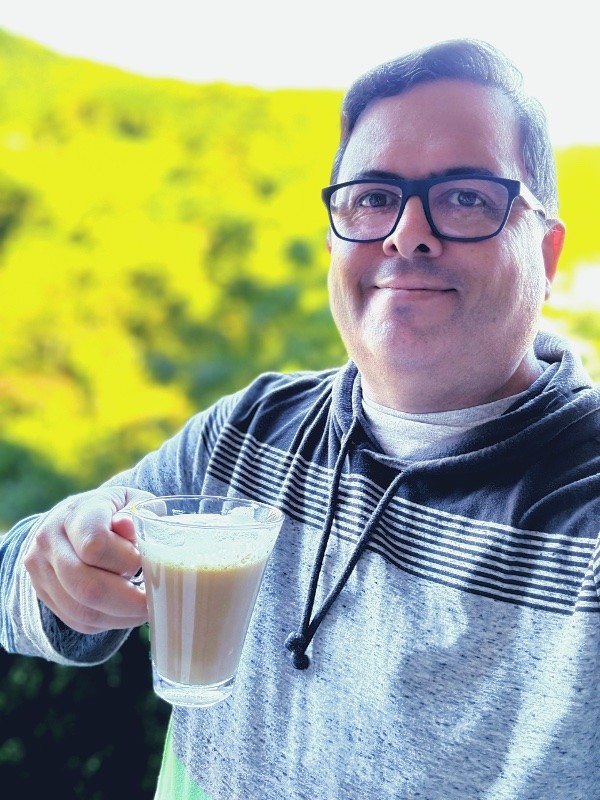 Selfie minha de casaco, sorrindo para a câmera, segurando uma xícara transparente contendo café com leite, com a mata iluminada de sol ao fundo.