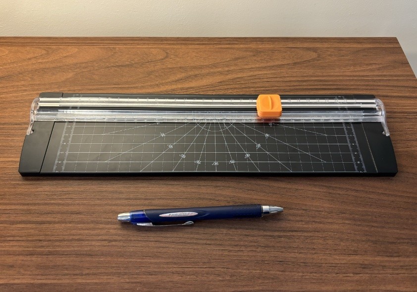 Uma guilhotina para papeis, formato A4, junto a uma caneta para permitir a escala.