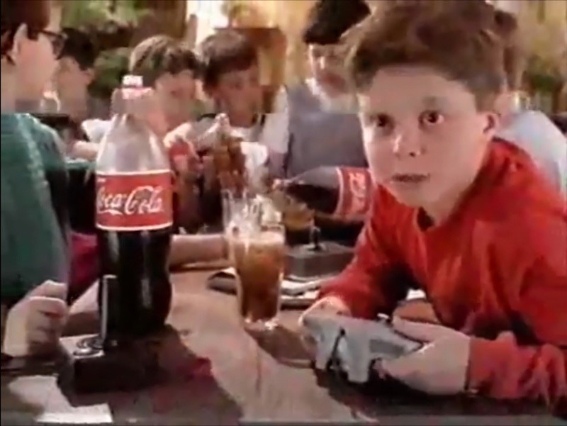 Criança segura um controle de videogame em frente a uma garrafa de Coca-Cola, em comercial de 1999.