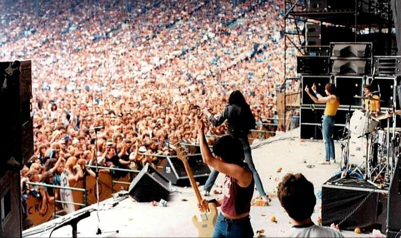 Ramones no palco, com Johnny, sem camisa, erguendo as duas mãos, com os dedos do meio em riste.