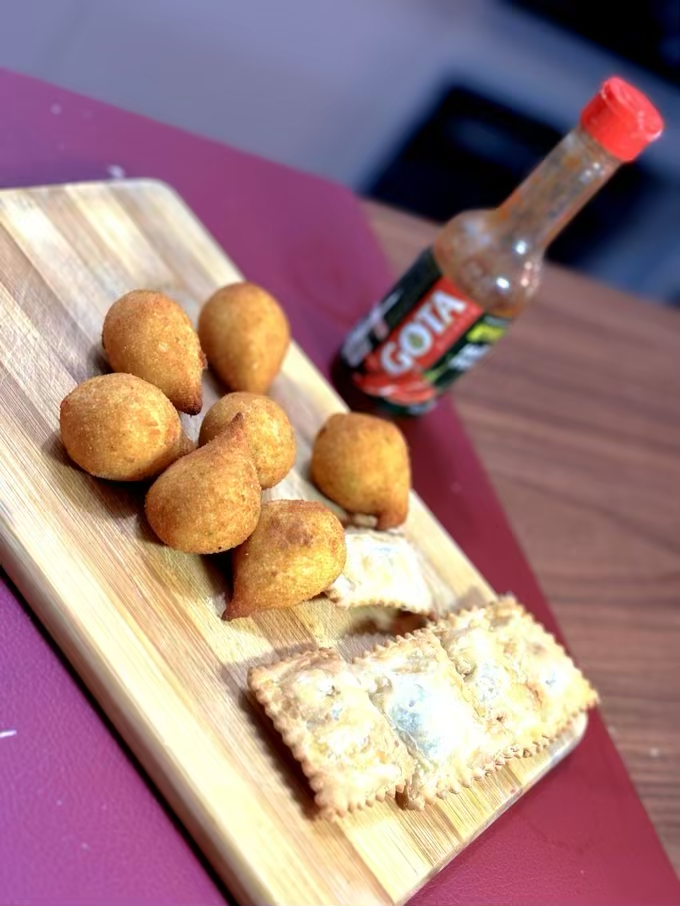 Uma tábua com 7 mini-cozinhas e 4 pasteizinhos, junto a uma embalagem de molho de pimenta marca Gota