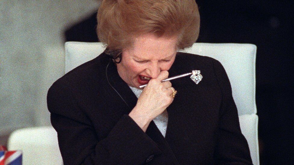 Thatcher, com expressão facial de desgosto, segura um lápis branco próximo à boca