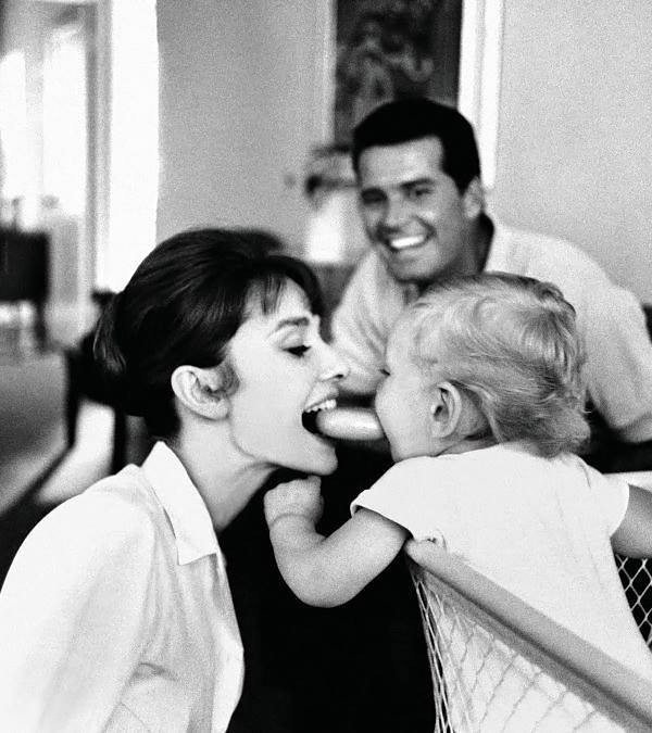 Em foto publicitária em preto e branco, o papai sorri ao fundo enquanto a mamãe, também sorridente, entrega com os dentes um objeto ao bebê, que o recebe com a boca