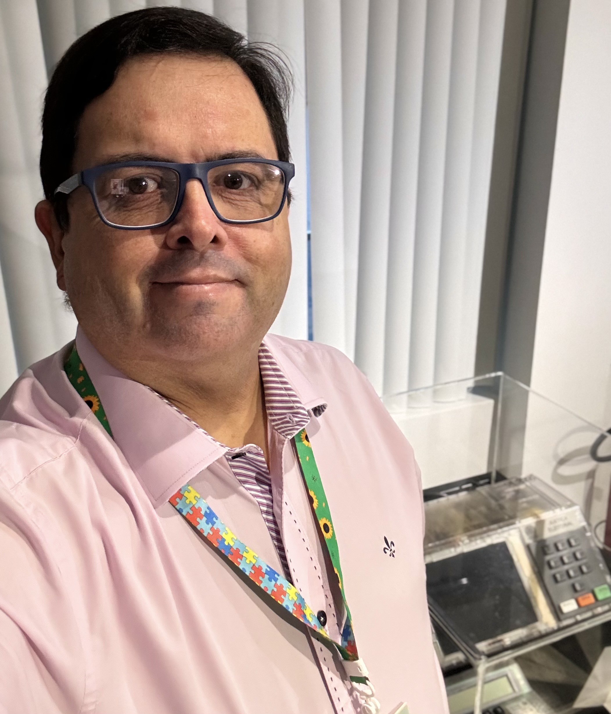 Foto minha olhando para a câmera, com camisa social rosa e cordão de crachá com símbolos de quebra-cabeça e de girassol, ao lado de uma urna eletrônica com gabinete transparente 