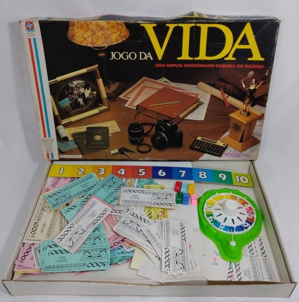 Caixa da versão clássica do Jogo da Vida, aberta e mostrando a roleta, as cédulas e os carrinhos que indicam a posição do jogador no tabuleiro.