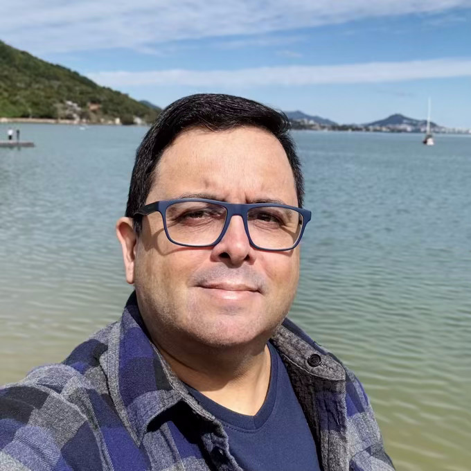 Foto minha olhando para a câmera, de jaqueta azul grunge, com a baía norte de Florianópolis ao fundo