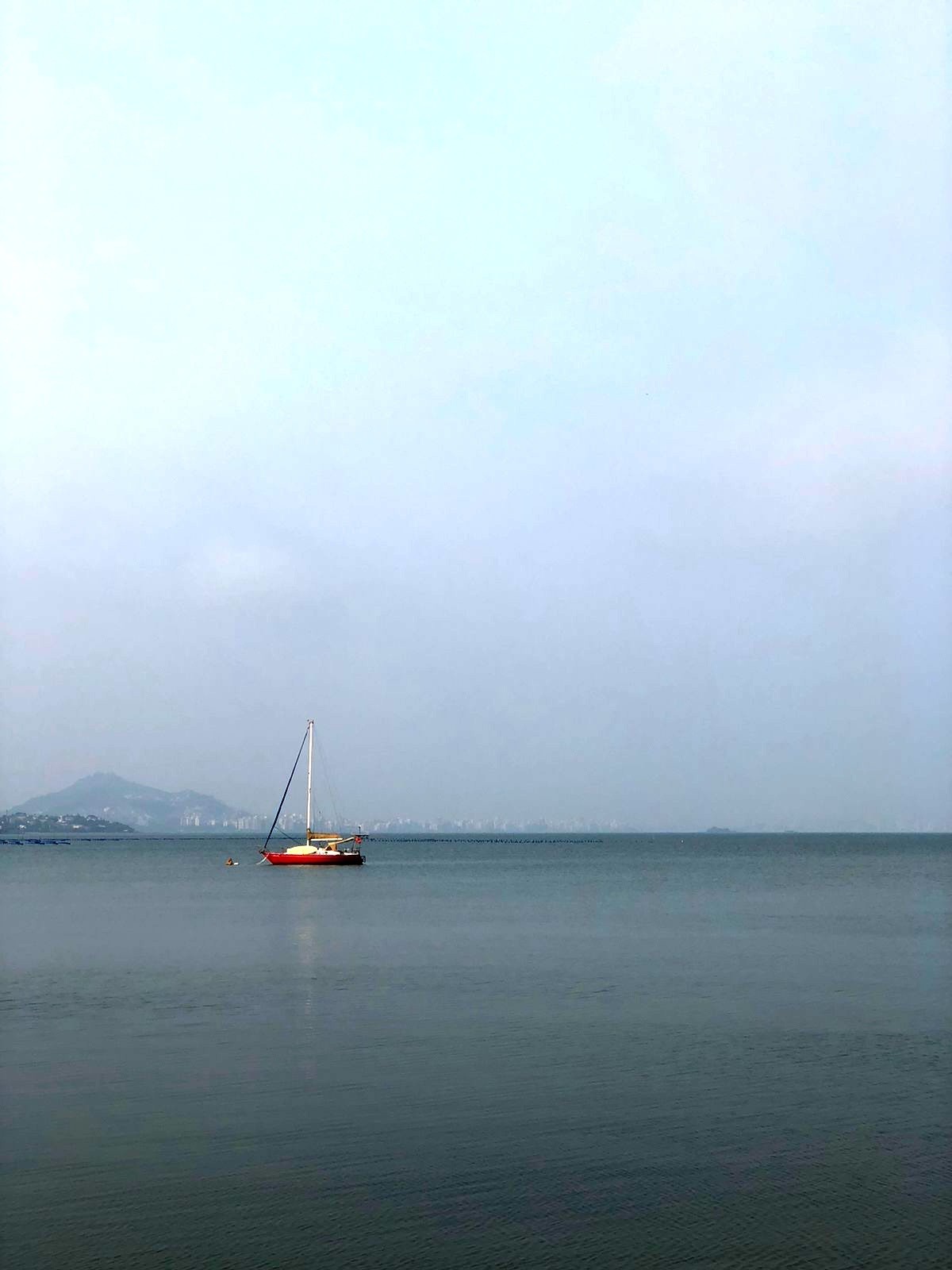 Foto de um veleiro com casco vermelho, fundeado próximo à costa, em uma baía de águas mansas, com o horizonte ao fundo, e a Avenida Beira-Mar Norte parcialmente visível em meio à névoa.